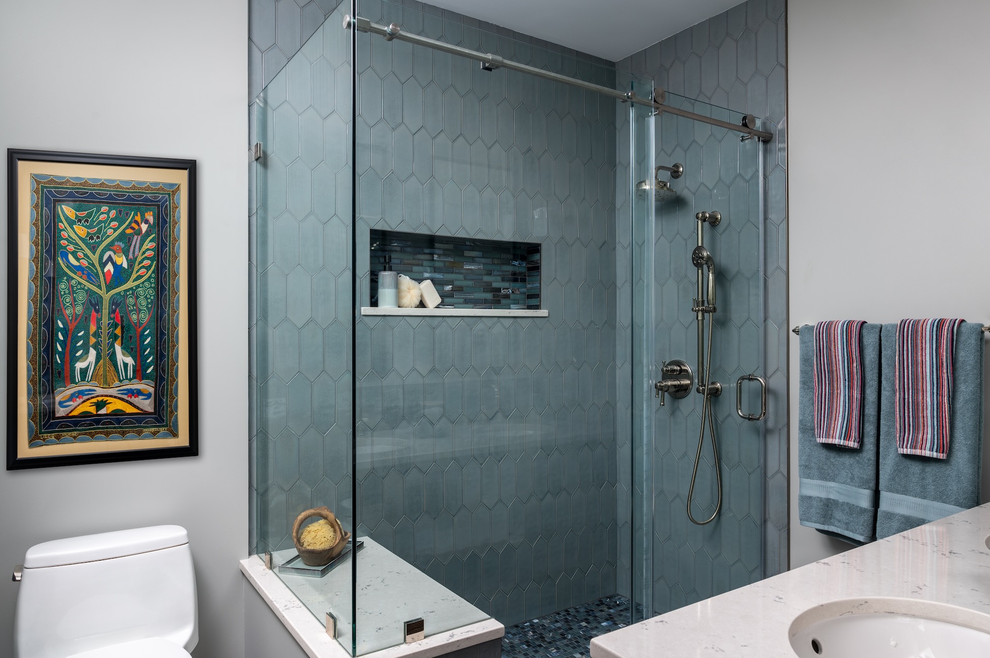 Renovated glass-enclosed shower, quartz-topped shower bench, blue-gray picket-style wall tile, niche lined with mosaic bar tile.