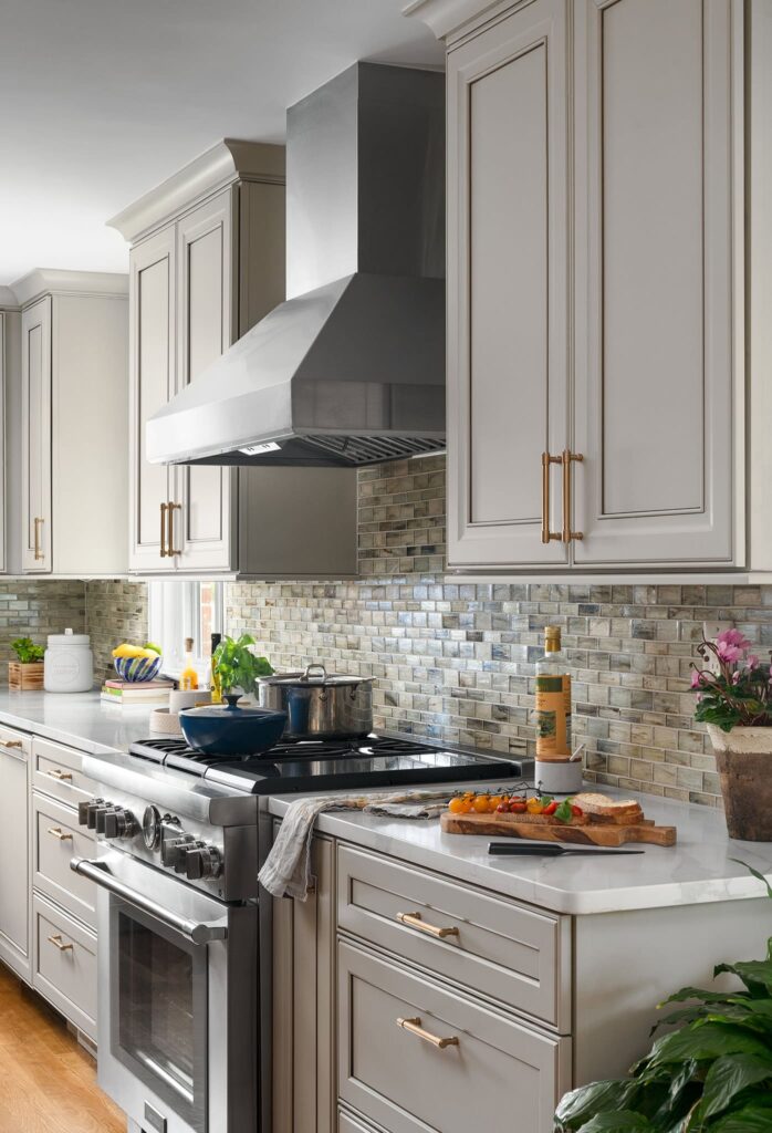 Renovated transitional kitchen with stainless steel Thermador range, Zephyr hood, gray-painted cabinetry, glass mosaic tile backsplash, brass hardware.