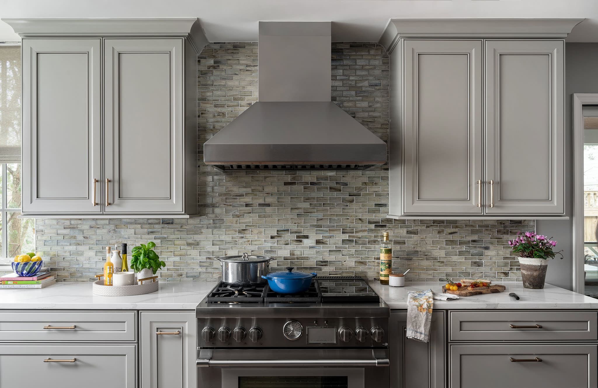 Kitchen range wall with stainless steel Thermador range, Zephyr hood, gray-painted cabinets, glass mosaic backsplash, brass hardware.
