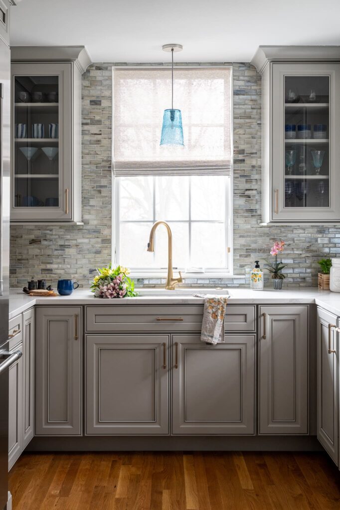 Kitchen sink wall with casement window, blue glass pendant, mosaic glass tile backsplash and gray-painted cabinetry, brass faucet and hardware.