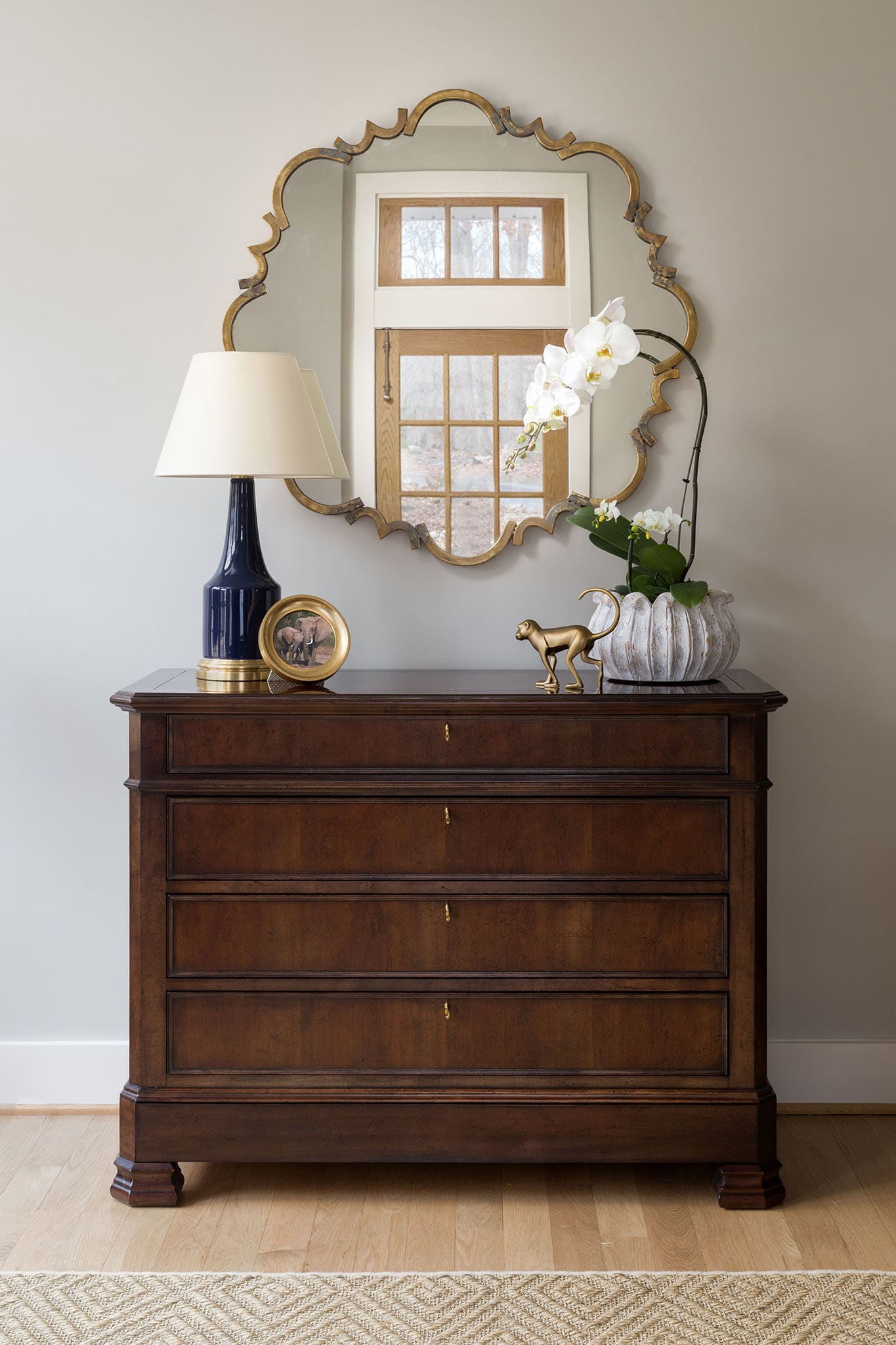 A dark wooden dresser with four drawers is topped with a lamp, framed photo, gold horse figurine, and a vase of white orchids. A decorative mirror hangs above, reflecting a window.