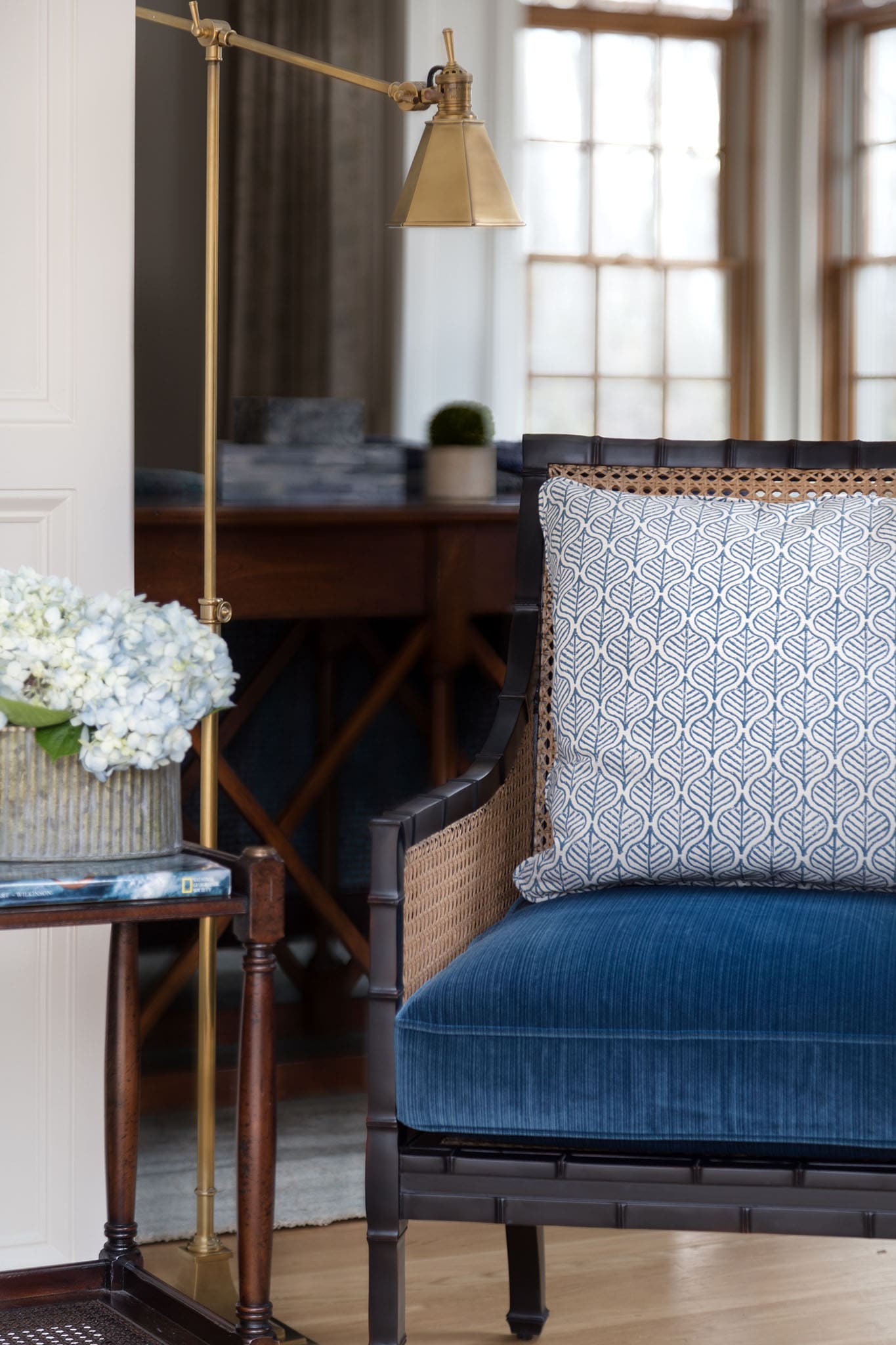 A wooden armchair with blue velvet seat and patterned pillow sits next to a table with white flowers and a brass floor lamp in a bright, windowed room.A wooden armchair with blue velvet seat and patterned pillow sits next to a table with white flowers and a brass floor lamp in a bright, windowed room.