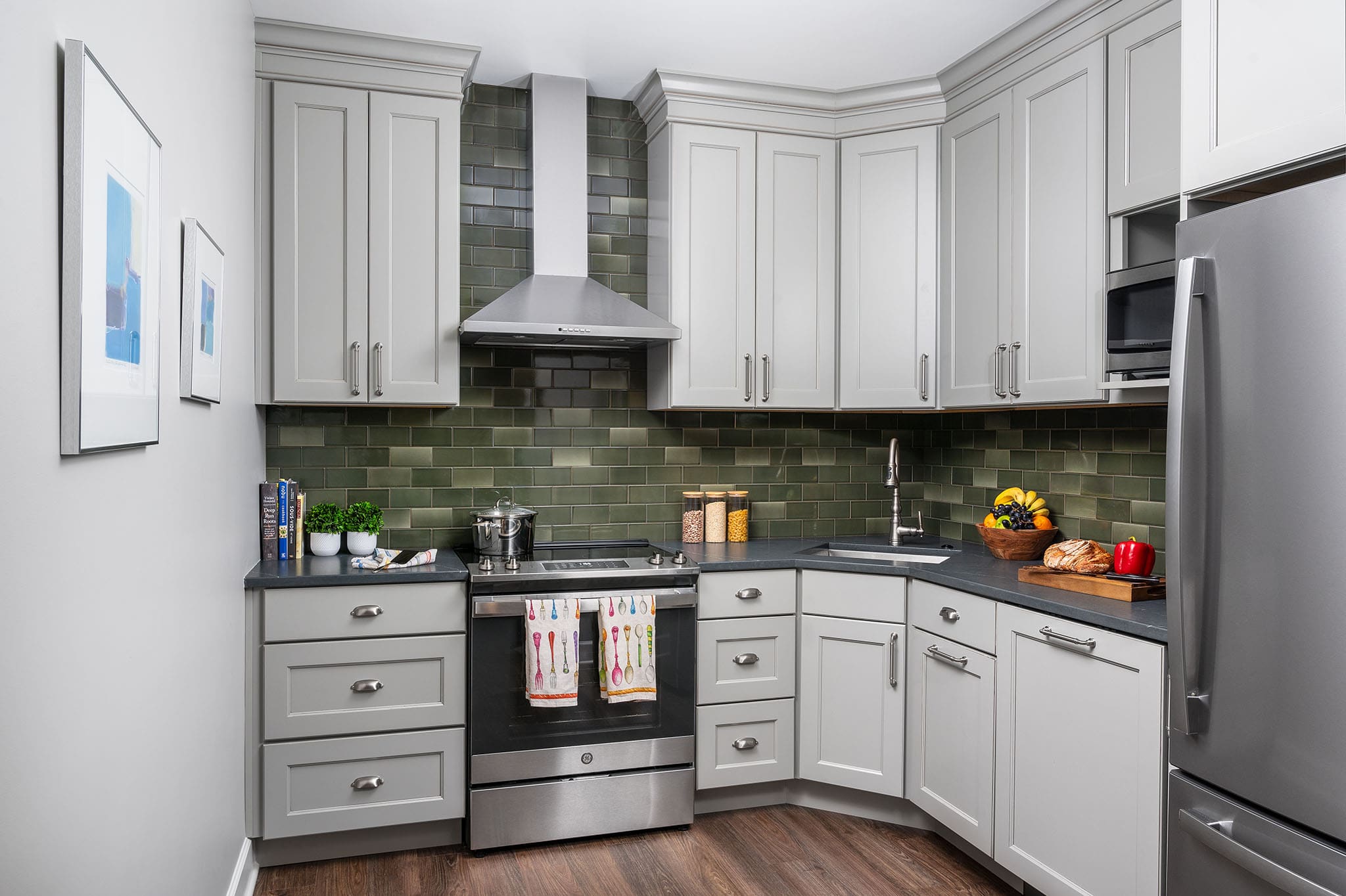 Modern kitchen with white cabinets and green subway tile backsplash. Stainless steel appliances and countertops feature fruit, bread, and decor. Warm, inviting tone.