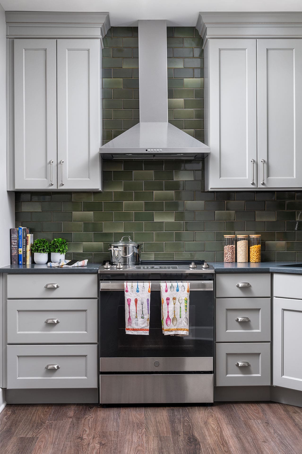 Modern kitchen with grey cabinets and a green tile backsplash. A sleek range and matching hood are centered. Cheerful towels hang on the oven, adding a vibrant touch.
