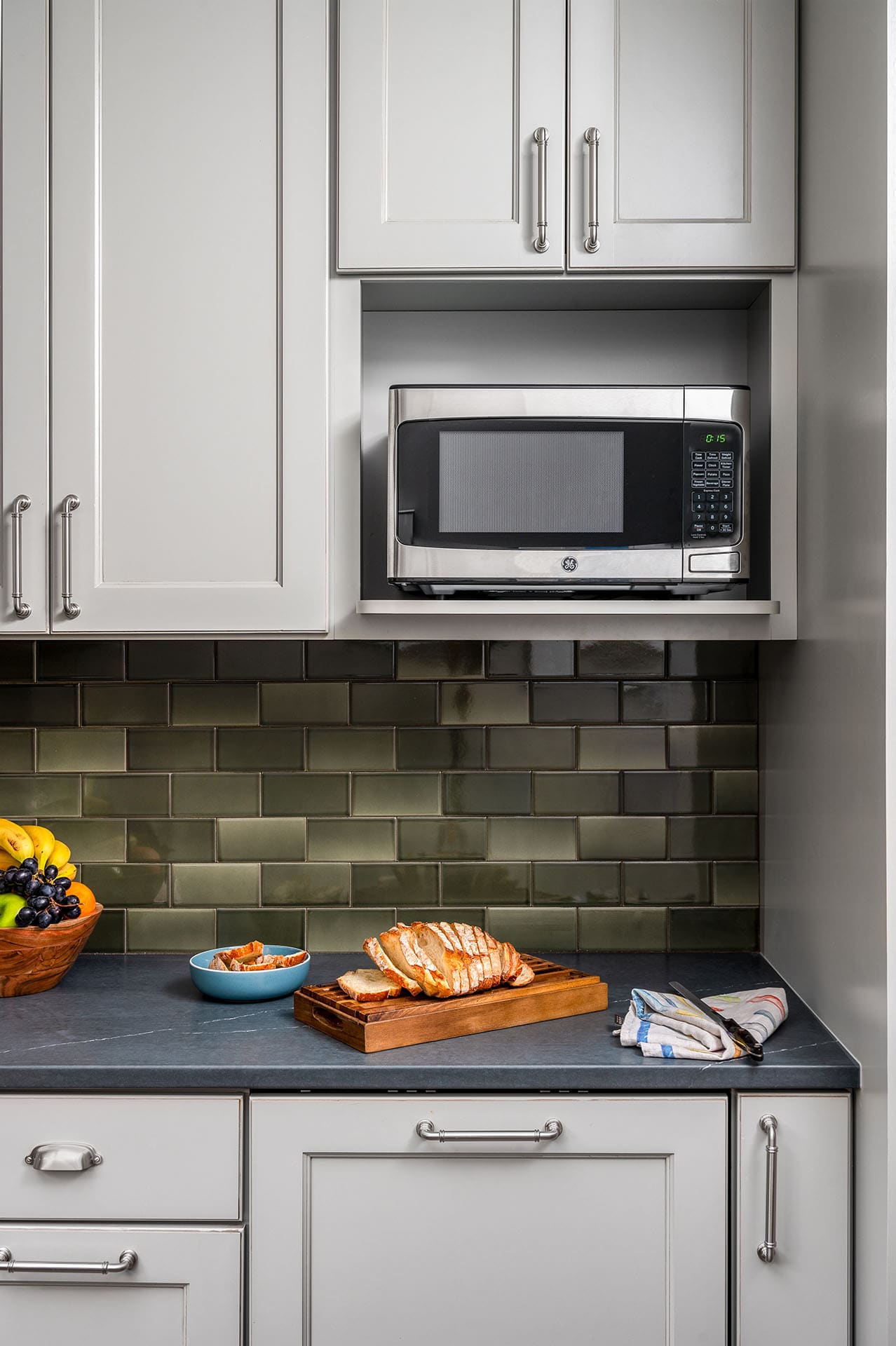 Modern kitchen countertop with green subway tile backsplash, featuring a gray microwave, sliced bread on a wooden board, fruit bowl, blue dish, and folded towels.