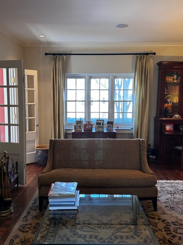 Living room with a brown sofa, glass coffee table, patterned rug, and a window showing trees; a wooden cabinet and framed photos are also visible.