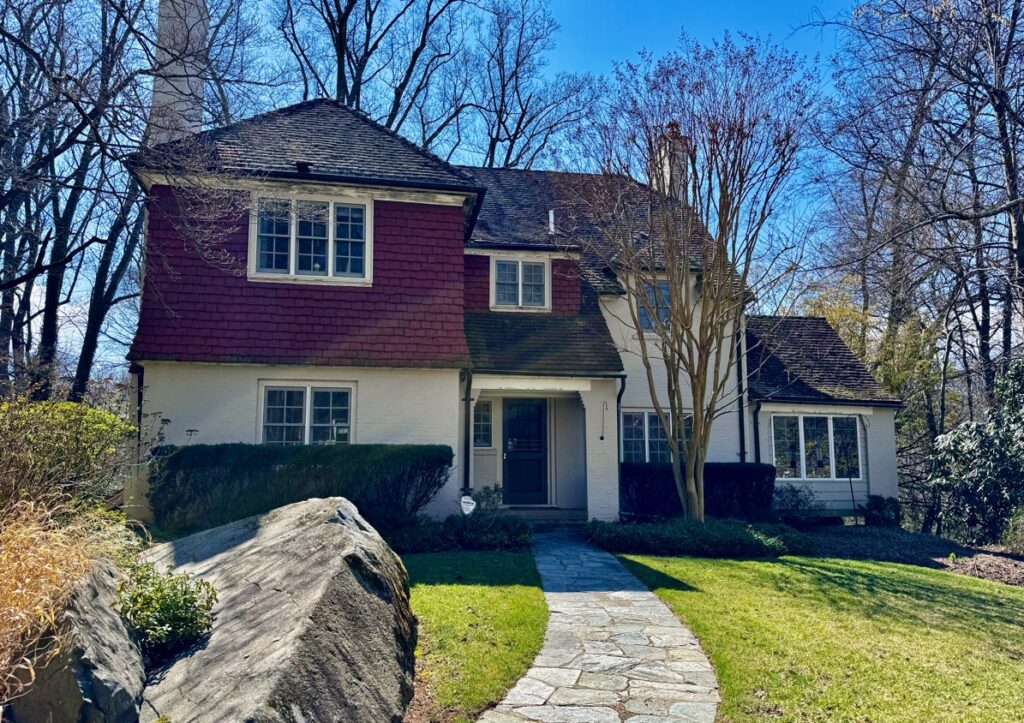 A two-story house with red shingles and white walls, surrounded by green grass, trees, and a stone pathway leading to the entrance.