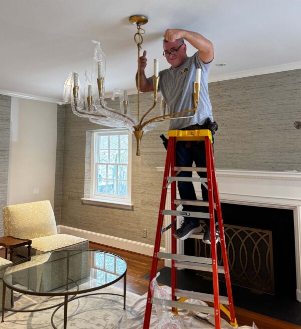 A man installs a golden chandelier while standing on a ladder in a bright living room. Decor includes a glass coffee table.