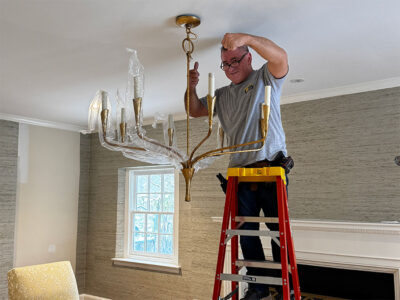 A man installs a gold chandelier on a ladder in a stylish living room with neutral walls and a cozy atmosphere.