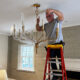 A man installs a gold chandelier on a ladder in a stylish living room with neutral walls and a cozy atmosphere.