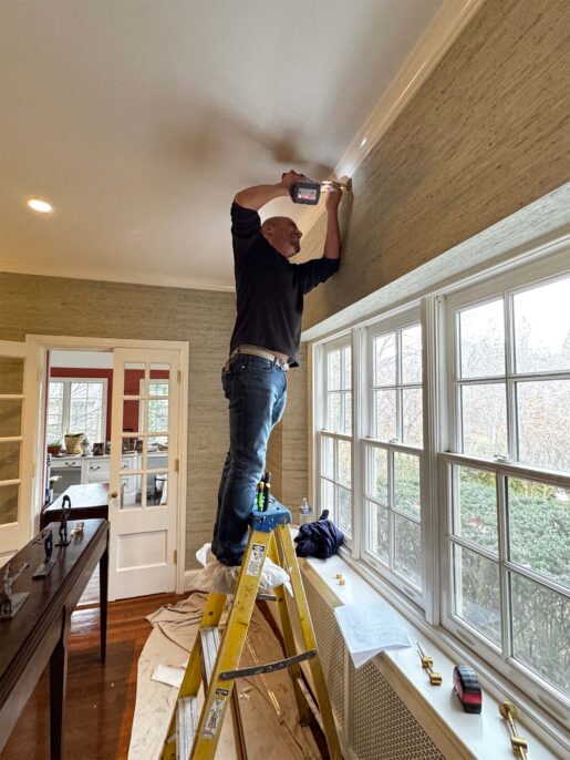 Man standing on a ladder, installing lighting fixtures in a home with bright windows.