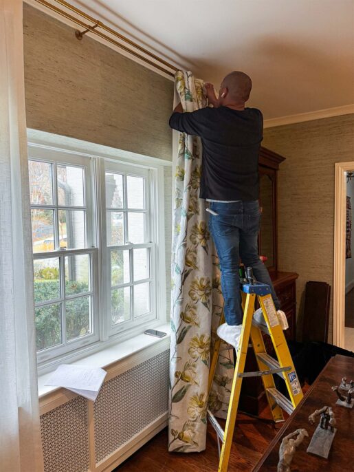 Person hanging floral curtains on a window while using a ladder, with notes and a phone nearby. Bright indoor setting.