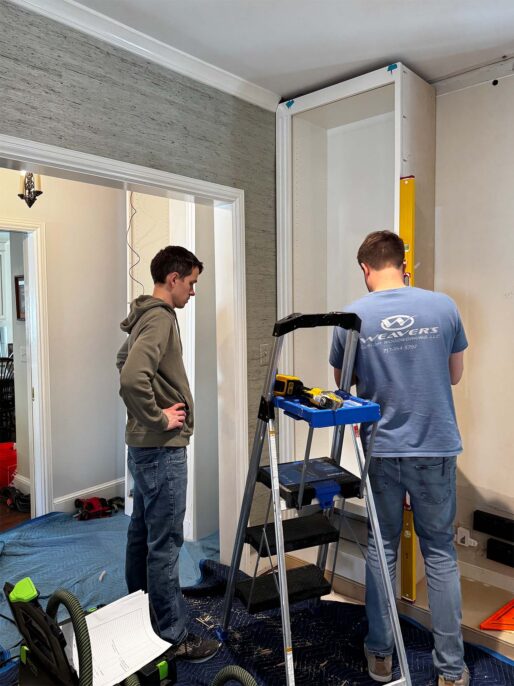 Two men installing a white shelving unit in a room, standing on a ladder with tools and protective coverings on the floor.