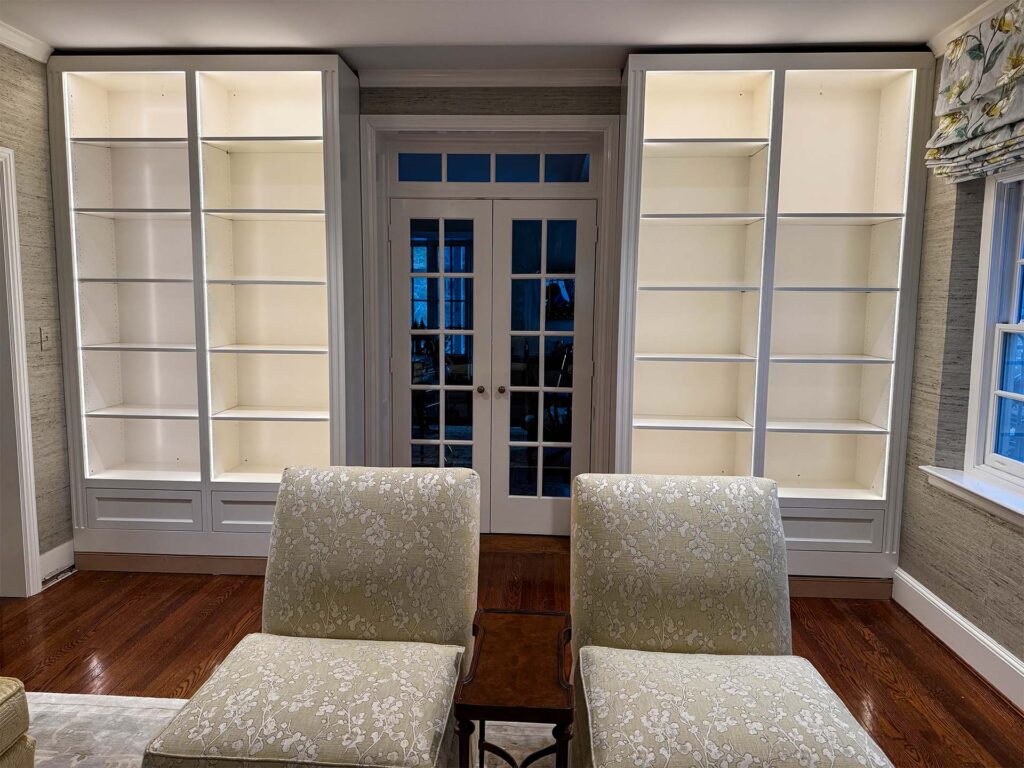 Bright, empty white bookshelves with decorative chairs in a cozy living room. Natural light illuminating the space.
