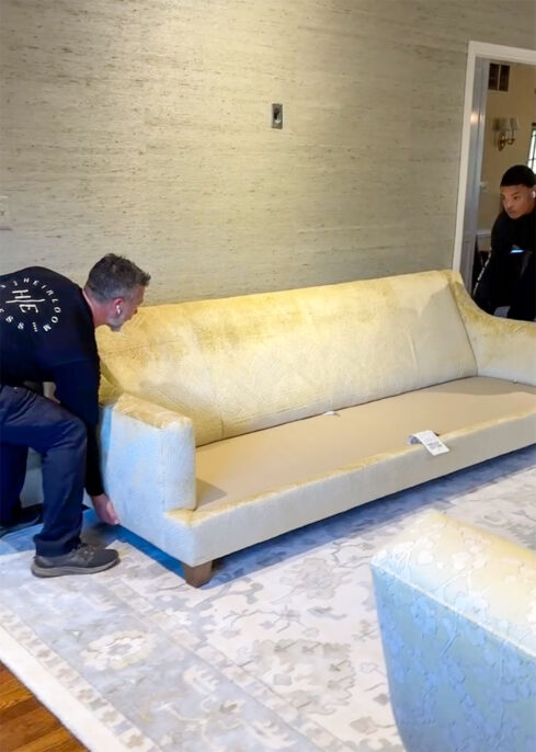 Two men carefully move a stylish gold sofa into a modern living room with an elegant rug.