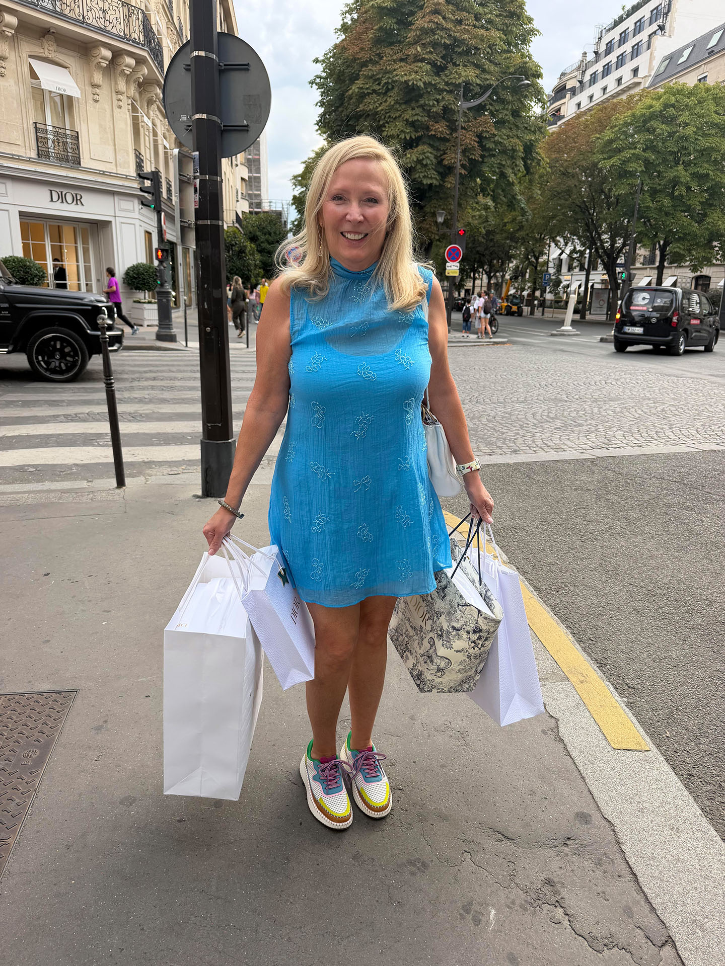 Andrea in a blue dress holding shopping bags on a bustling street near luxury boutiques in Paris.