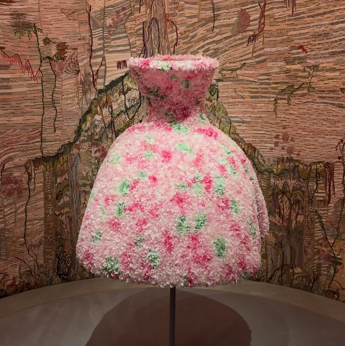 Colorful dress featuring pink and green textures, displayed against a detailed tapestry backdrop.